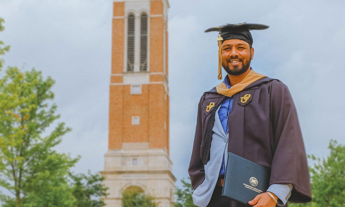 Man in cap and gown outside near brick bell tower.